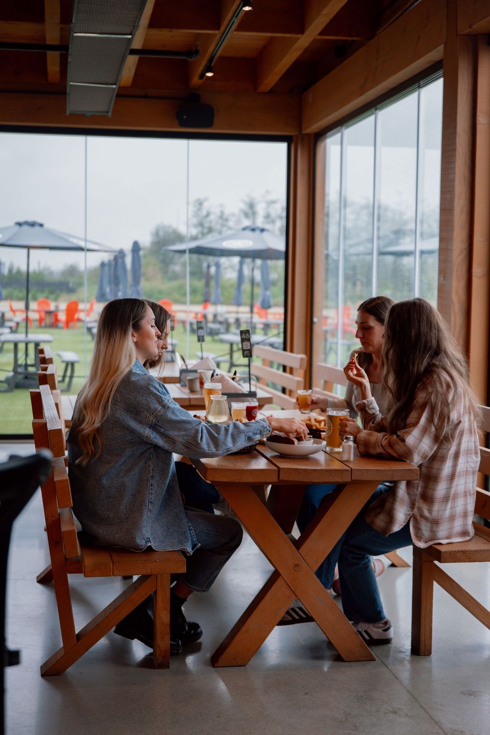 People dining at wooden picnic tables inside a restaurant with large windows overlooking a grassy outdoor area.