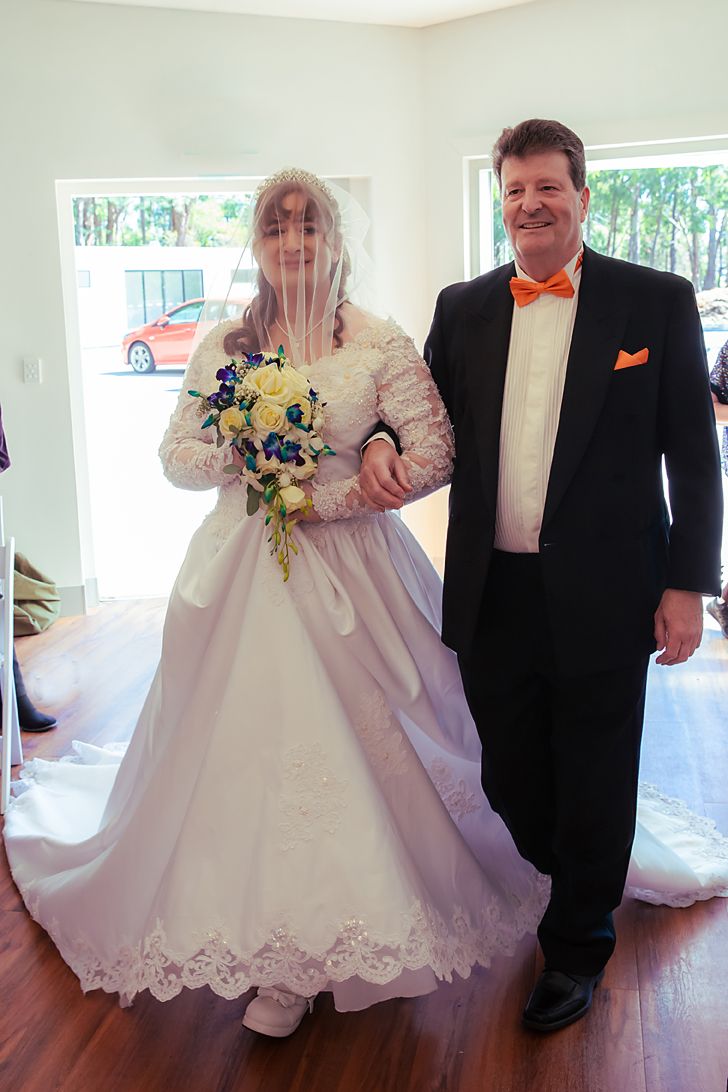 Wedding couple posing together on hardwood floor with bride wearing a white lace ballgown and groom in black tuxedo.