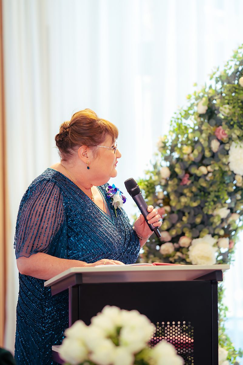 Person in a blue sequined dress speaks at a podium during a formal event with floral decorations in the background.