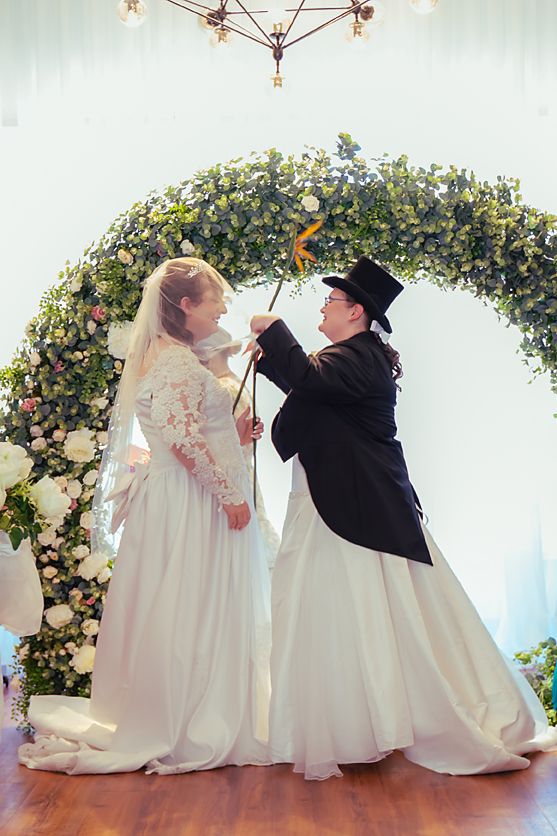 Two brides in matching white wedding gowns and floral arch share a romantic moment during a wedding ceremony photoshoot.