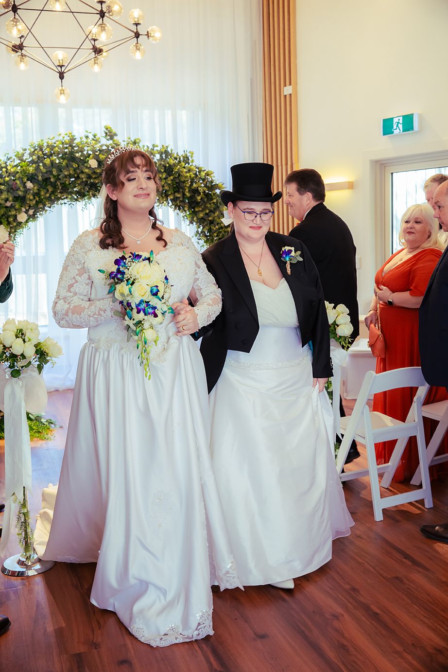 Wedding party members dance and celebrate on a polished wooden floor in an elegant room with chandeliers.