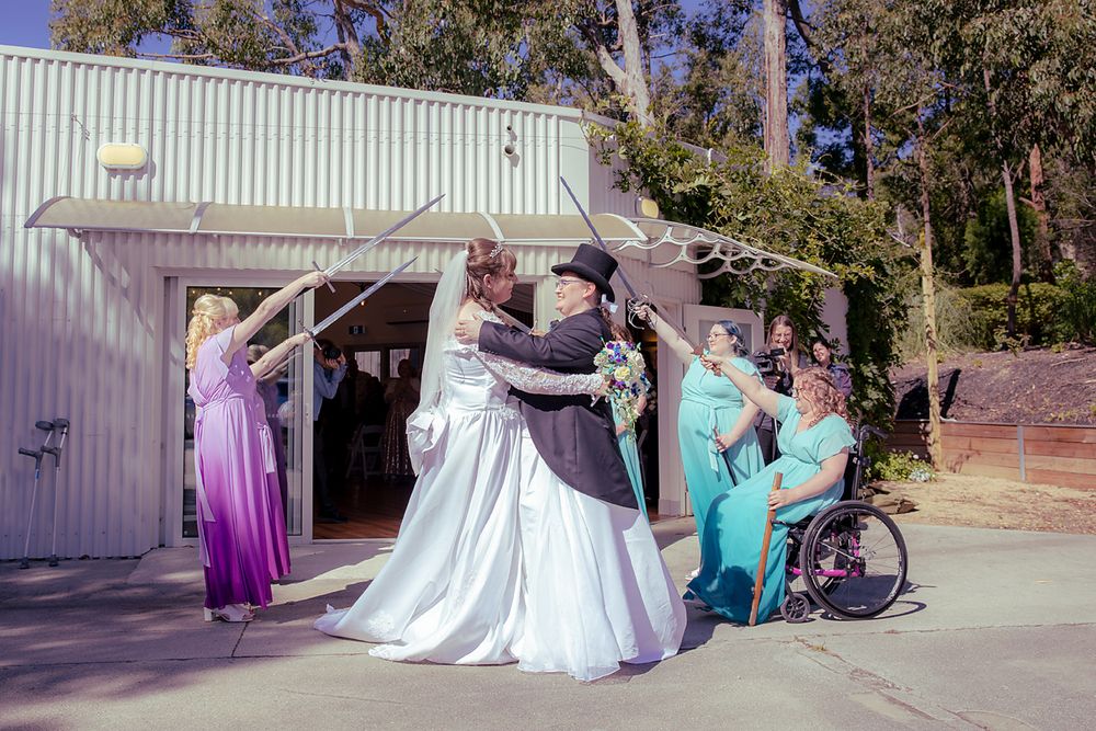 A group in formal attire including a white wedding gown and bridesmaid dresses in purple and teal gather outside a building.