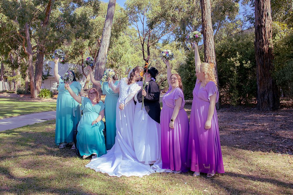 Wedding party wearing coordinated blue and purple dresses pose together outdoors among tall trees in a park setting.