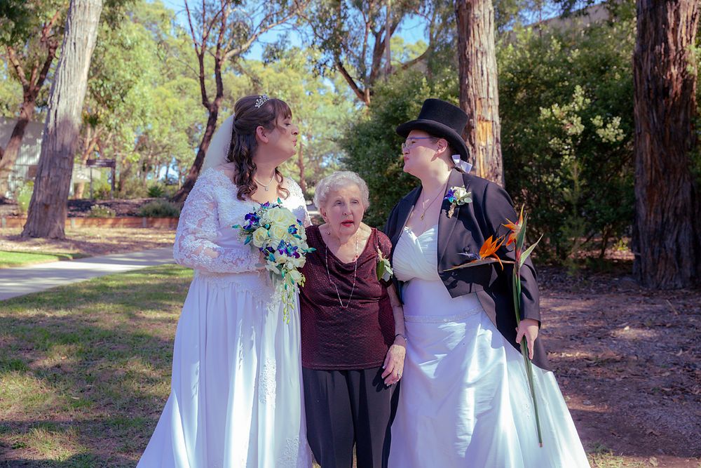 A romantic outdoor wedding ceremony takes place in a wooded area with tall pine trees in the background.