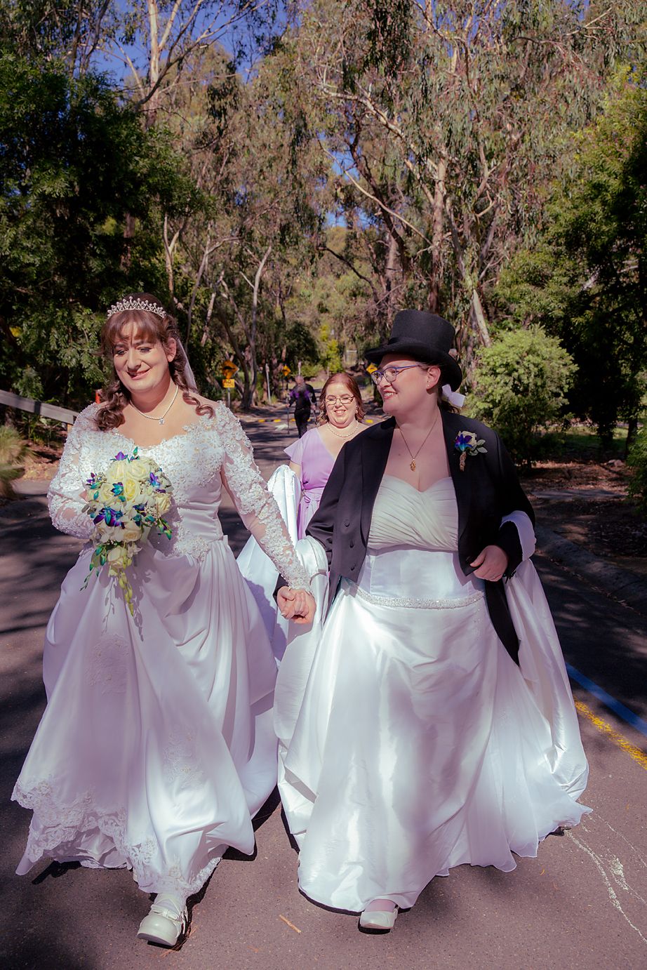 Two people in flowing white wedding dresses walk hand in hand outdoors, one wearing a black jacket and hat.
