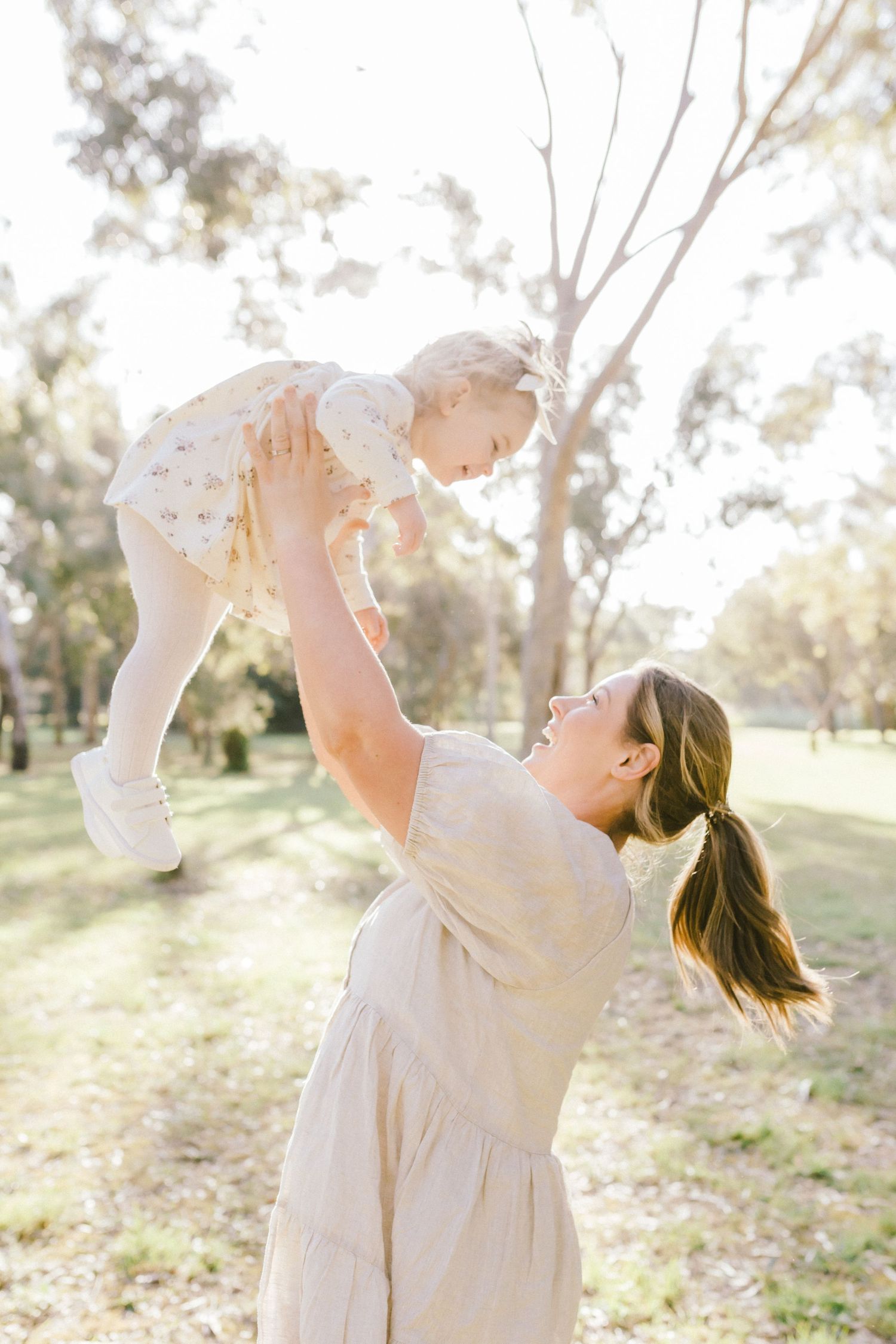 Parent and baby in white dresses play together in a sunlit outdoor setting with trees in the background.