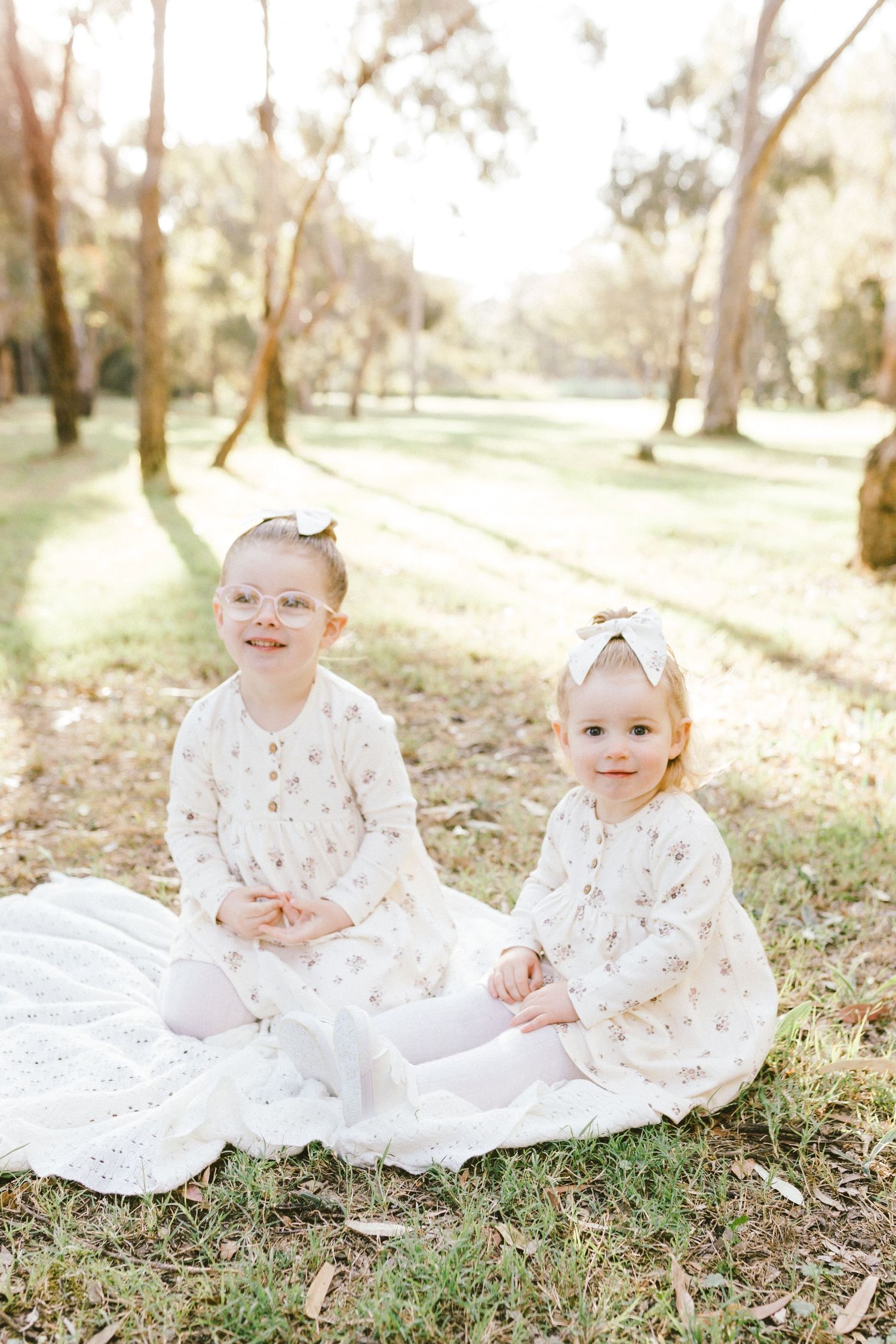 Infant in white lace dress sitting on grass in sunlit woodland setting with dreamy golden light filtering through trees.