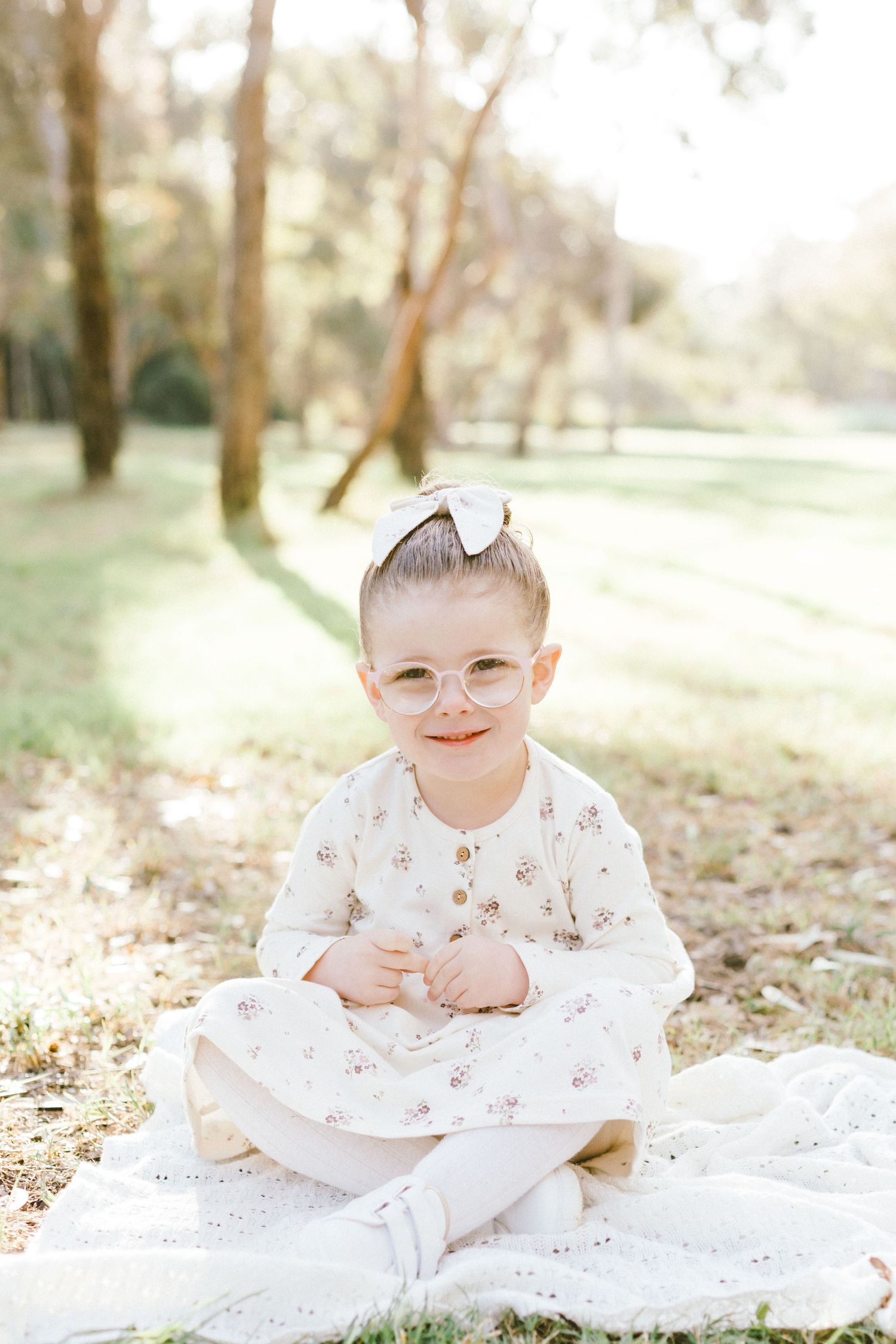 Baby wearing floral onesie lies on white blanket outdoors in soft natural lighting at a park.