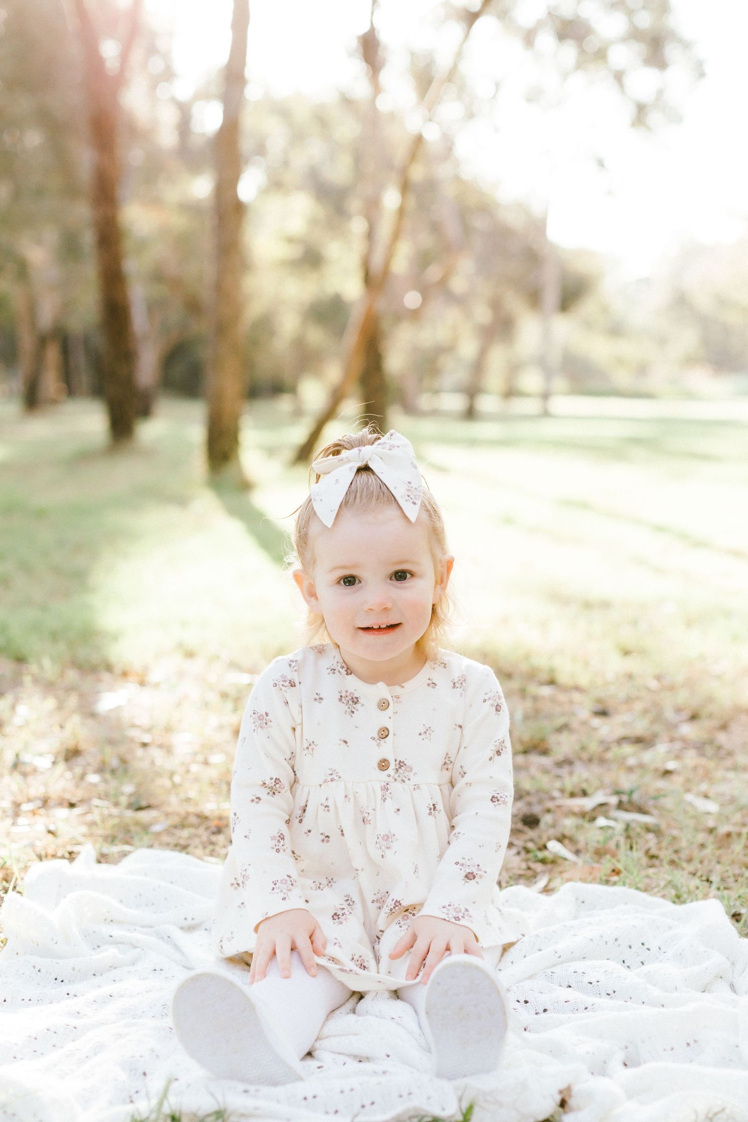 A small child wearing a white floral dress and bow sits on a blanket in a sunlit woodland setting.