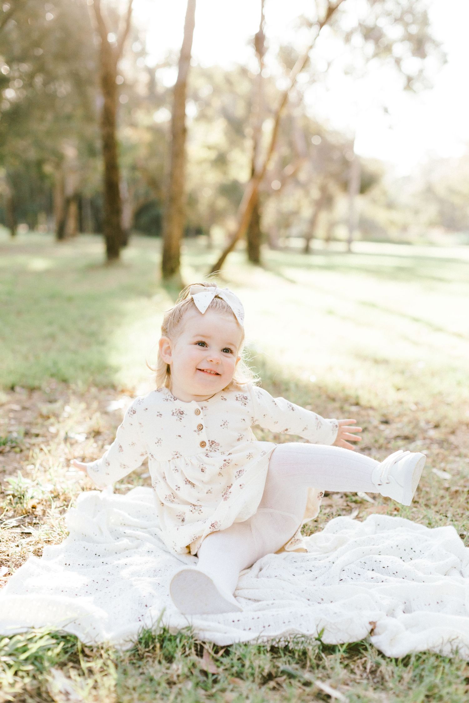 Small child in white lace dress lays on blanket outdoors in sunlit nature setting with soft bokeh background.