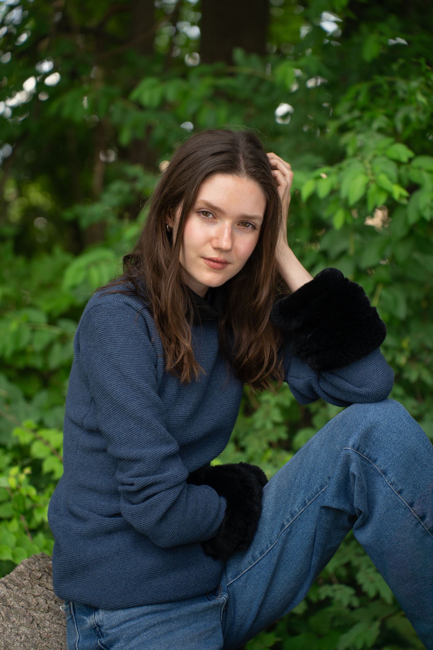 Client in blue sweater and jeans posing casually against green foliage background.