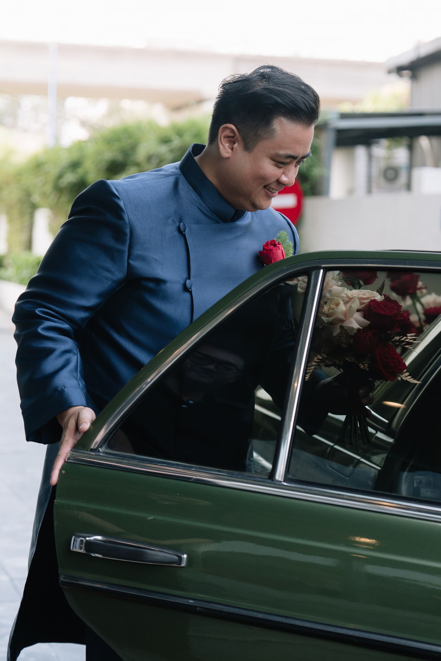Groom in blue ao dai suit with red rose boutonniere opening door of vintage green car at wedding.