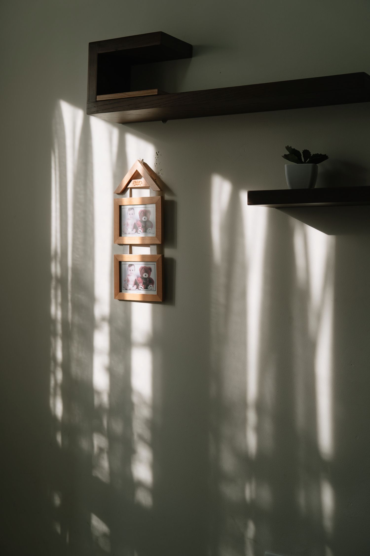 Wooden house-shaped photo frame hangs on green wall with sunlight shadows beside dark floating shelves.