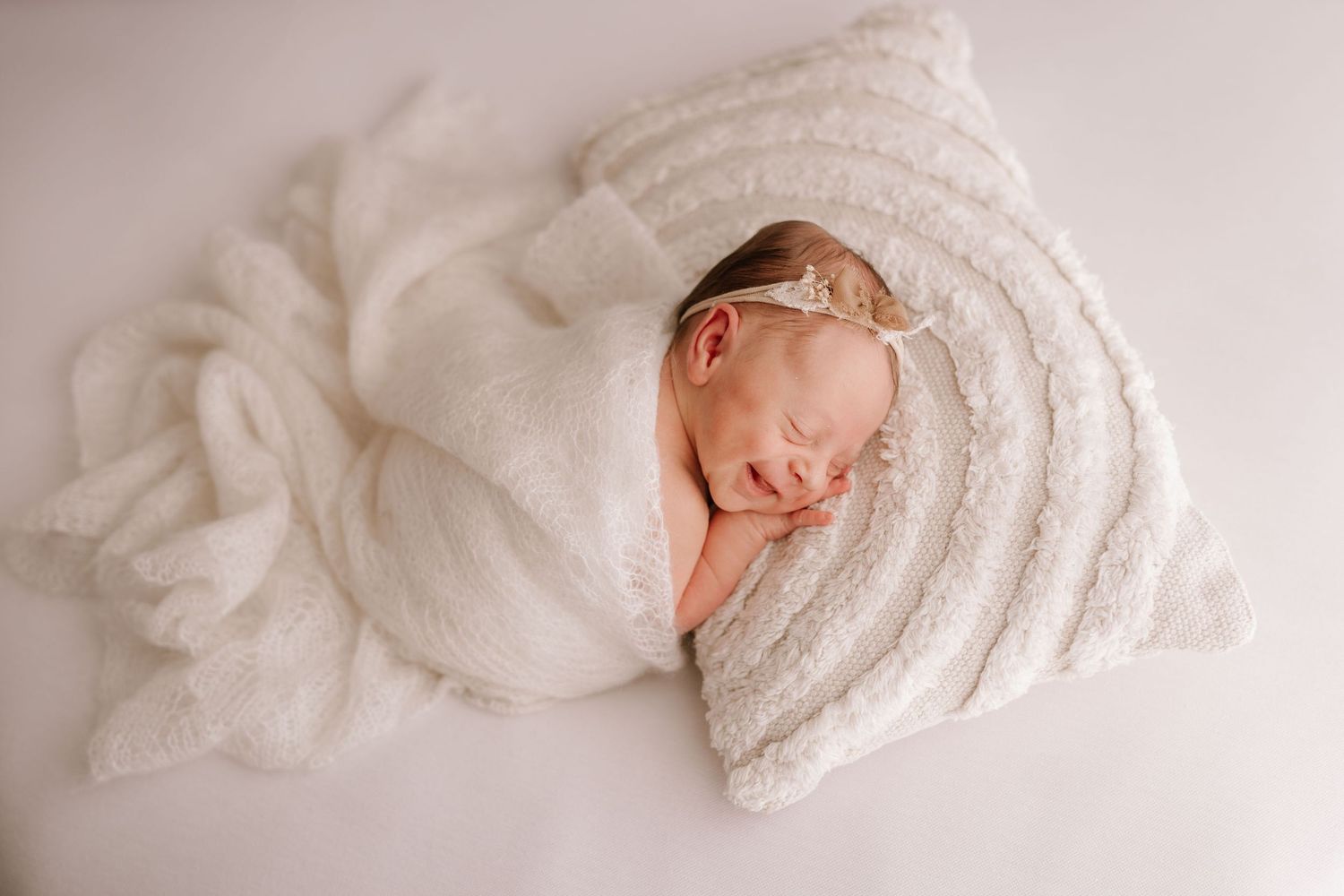 Newborn baby girl swaddled in white wrap sleeping on textured blanket with delicate floral headband.