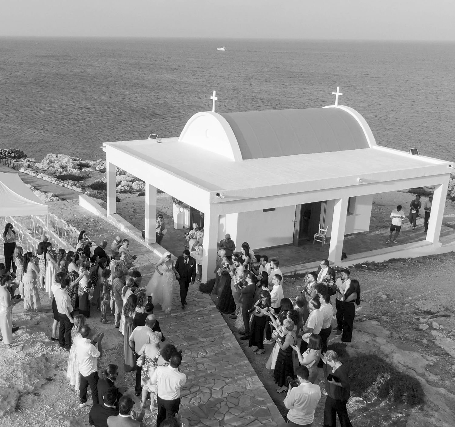 A crowd gathers outside a white domed Greek Orthodox church building overlooking the Mediterranean Sea in black and white.