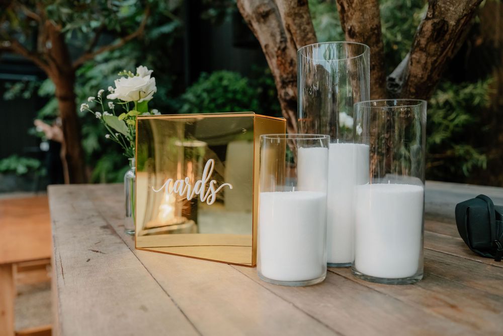 White candles and a gold metallic box arranged on a wooden table in an outdoor setting.