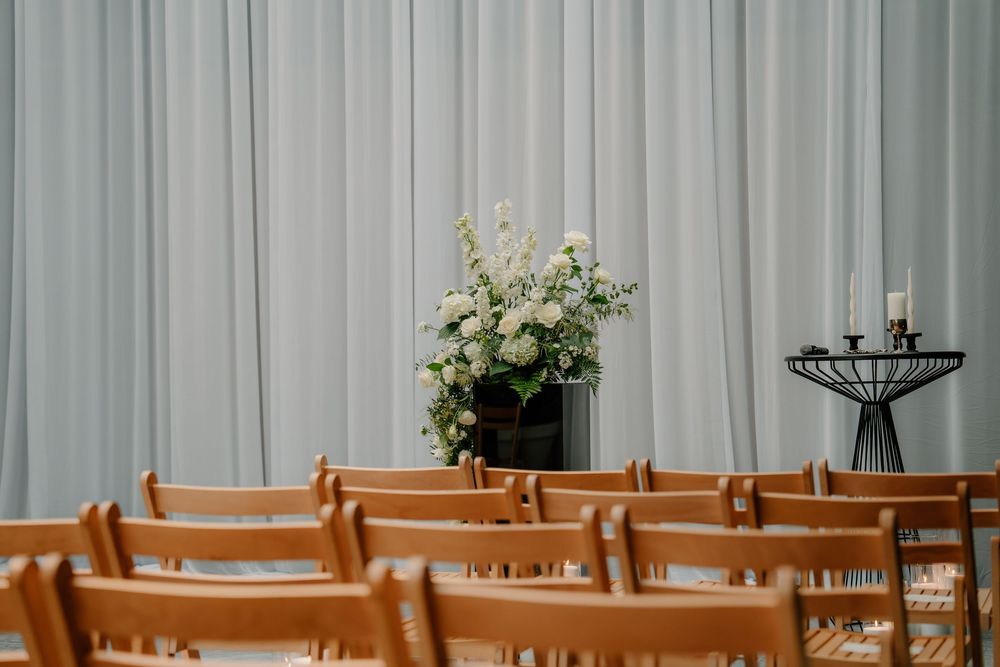 Wooden chairs lined up facing a floral arrangement and candlesticks against a white curtain backdrop.