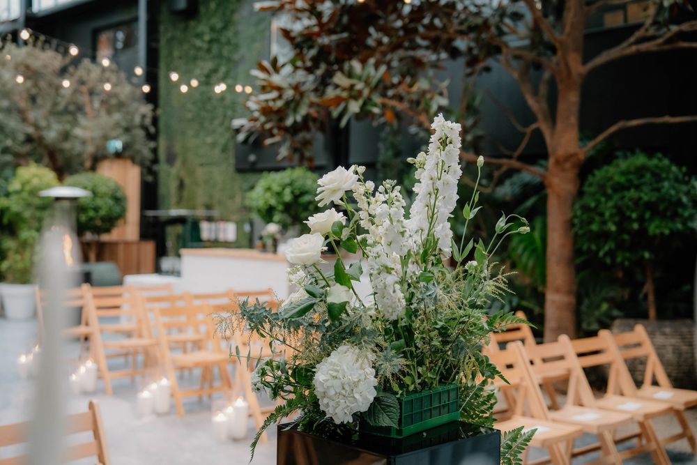 Elegant outdoor wedding reception setup with white floral centerpieces and wooden chairs arranged beneath tree canopy.