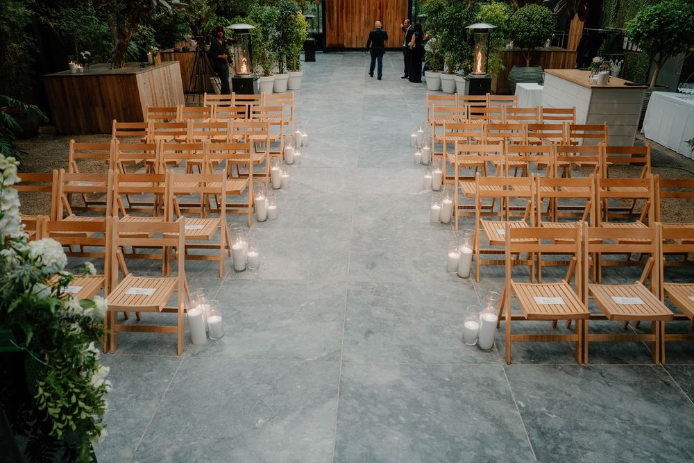Wide view of wooden ceremony chairs set up on stone patio with landscaping and traditional architecture.