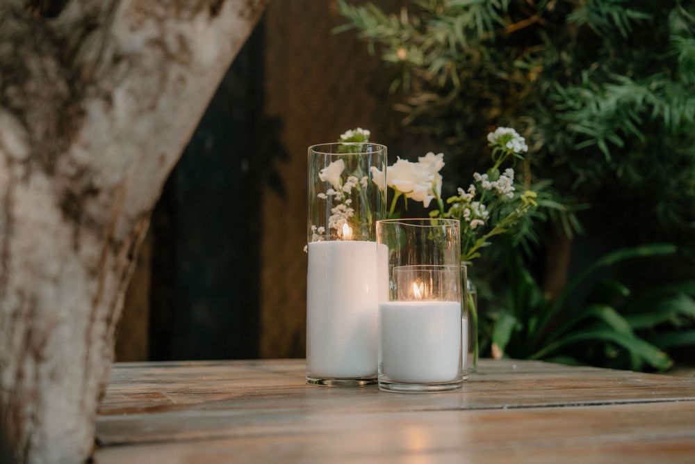 White pillar candles flicker in glass holders against lush greenery and rustic wooden window frames.