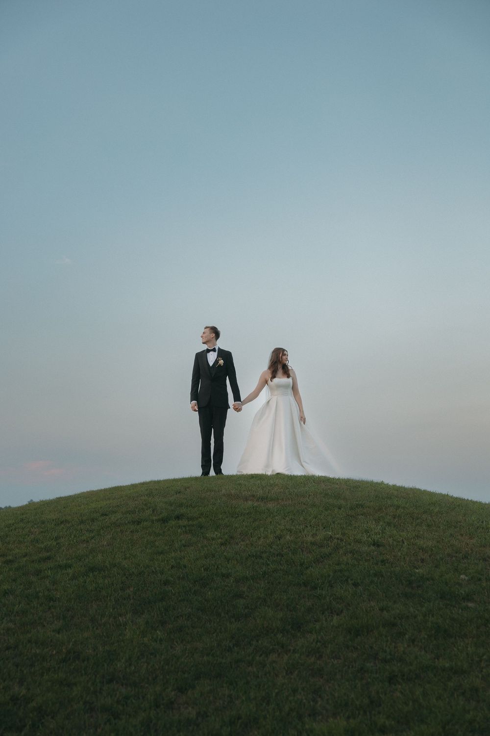 Wedding couple stands atop a grassy hill against a soft blue sky.