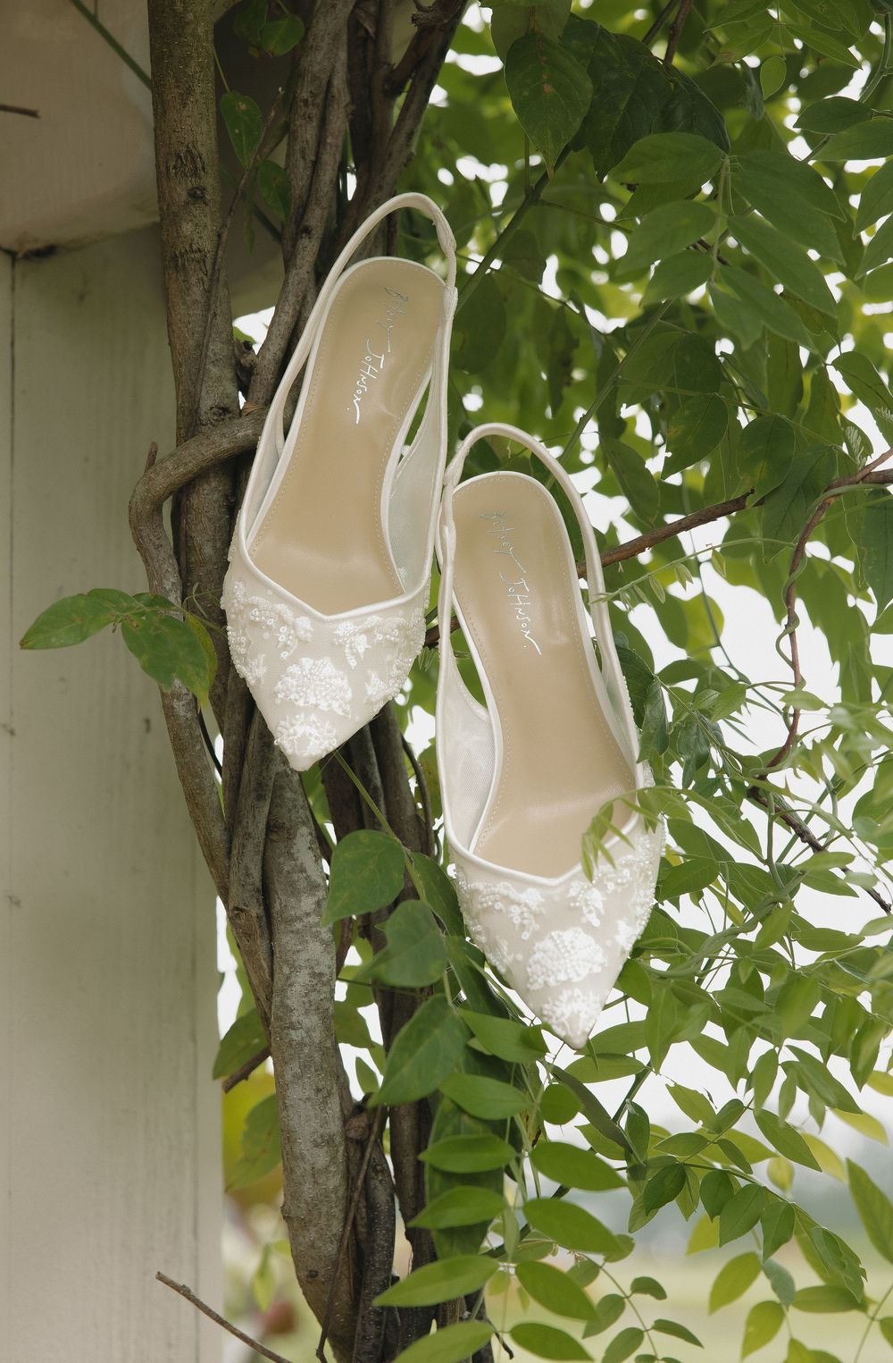 White sparkly bridal shoes hang from tree branches among green leaves.