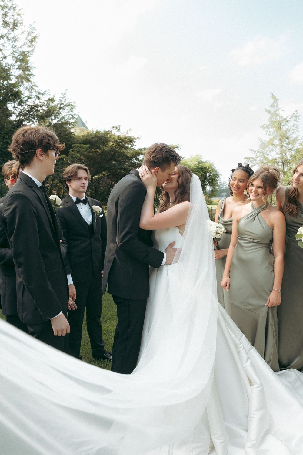 Wedding party laughs together while holding the bride's flowing veil outdoors.