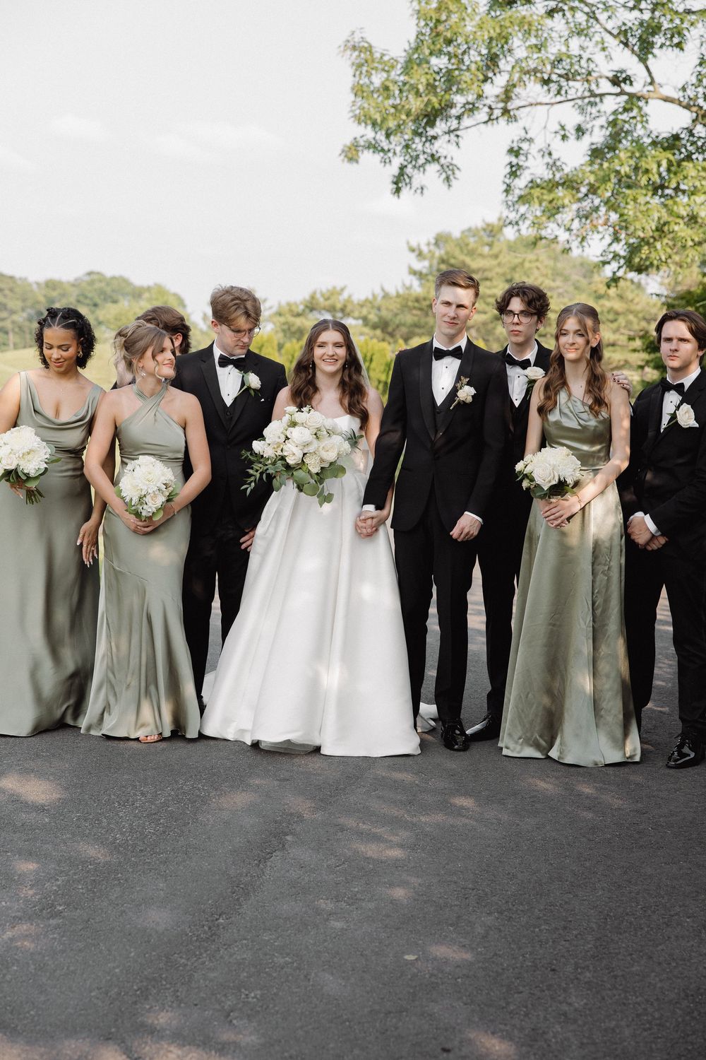 Wedding party poses in a line wearing black tuxedos and sage green bridesmaid dresses.