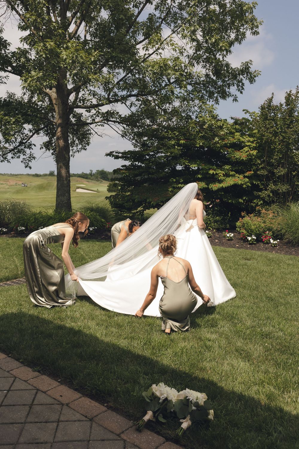 Wedding party helps arrange the bride's long train on grassy lawn.