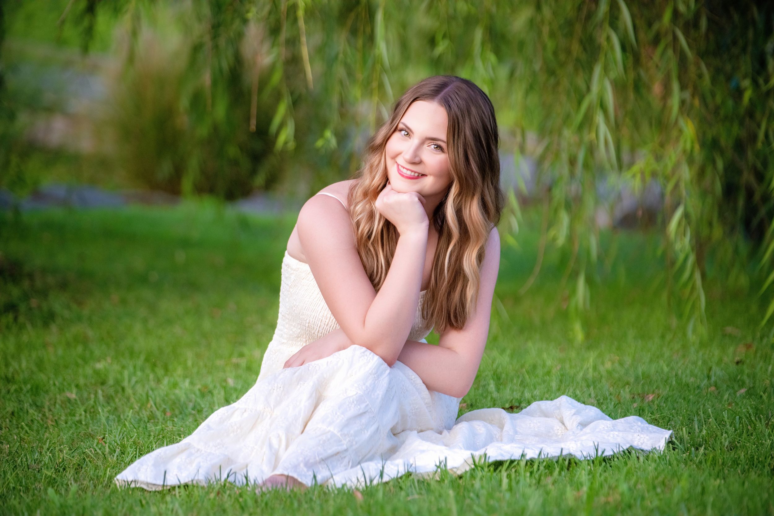 A person in a white dress reclines on lush green grass beneath drooping willow tree branches.