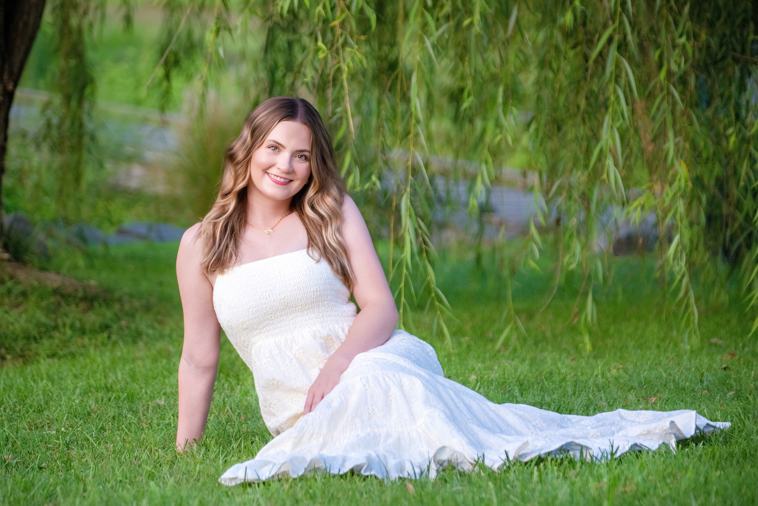 Figure in an ethereal white lace wedding dress poses gracefully under a weeping willow tree in a garden setting.
