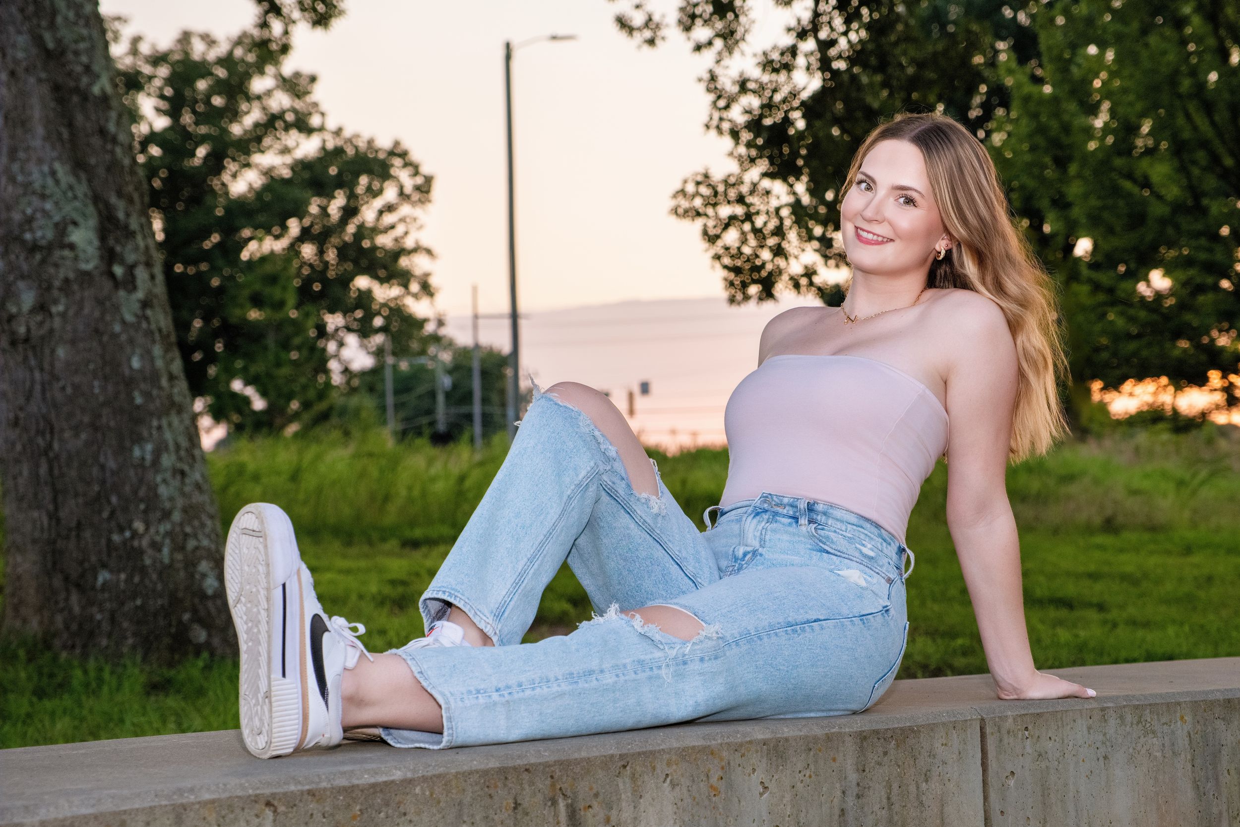 Person in light denim overalls and white sneakers reclines on concrete wall at sunset.