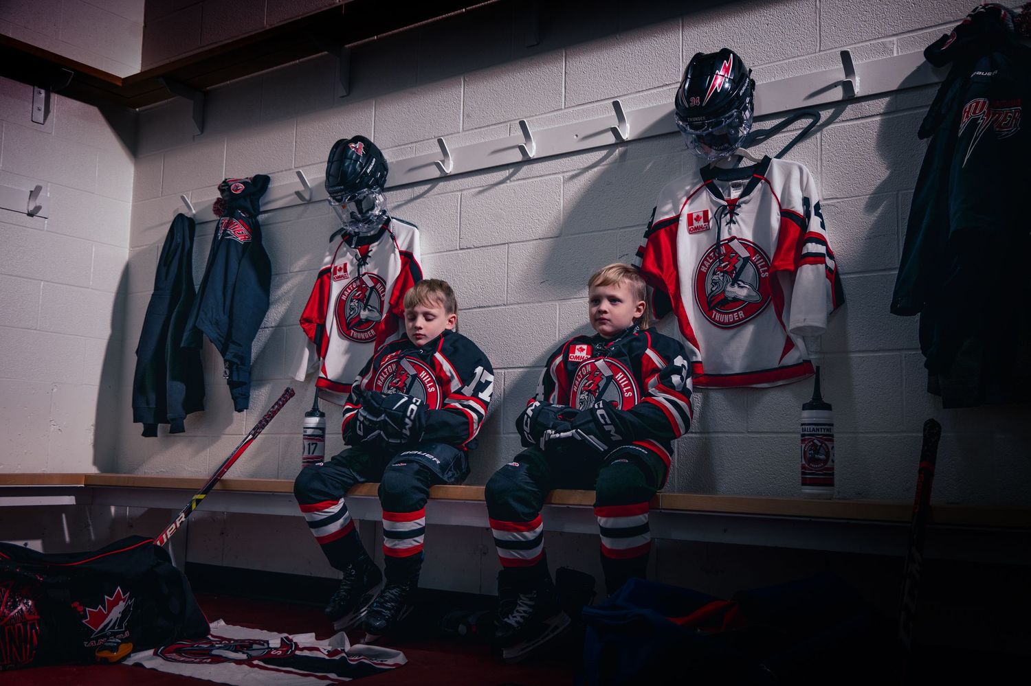 Two young hockey players sit in a locker room wearing red and white jerseys, helmets, and gloves ready for game.
