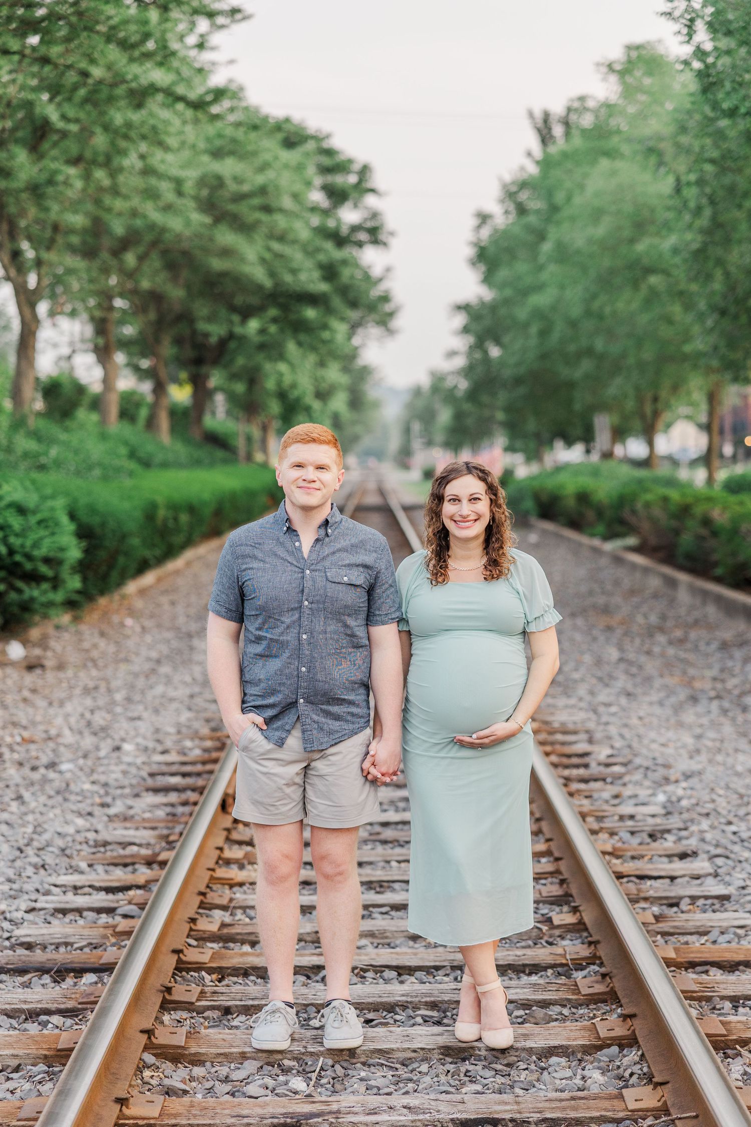 A couple stands together on railroad tracks surrounded by greenery during a maternity session.
