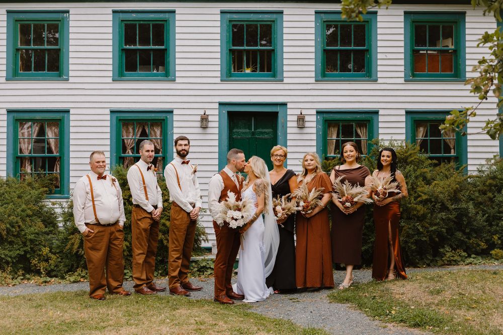 Wedding party poses together in front of white building with green trim windows.