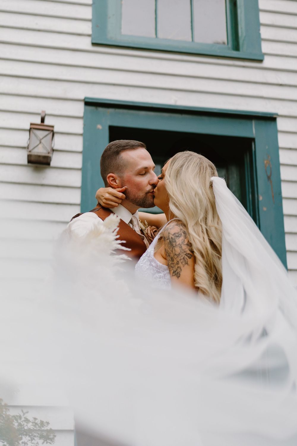 Romantic wedding photo through sheer white veil of a couple sharing a kiss against white building with green trim.