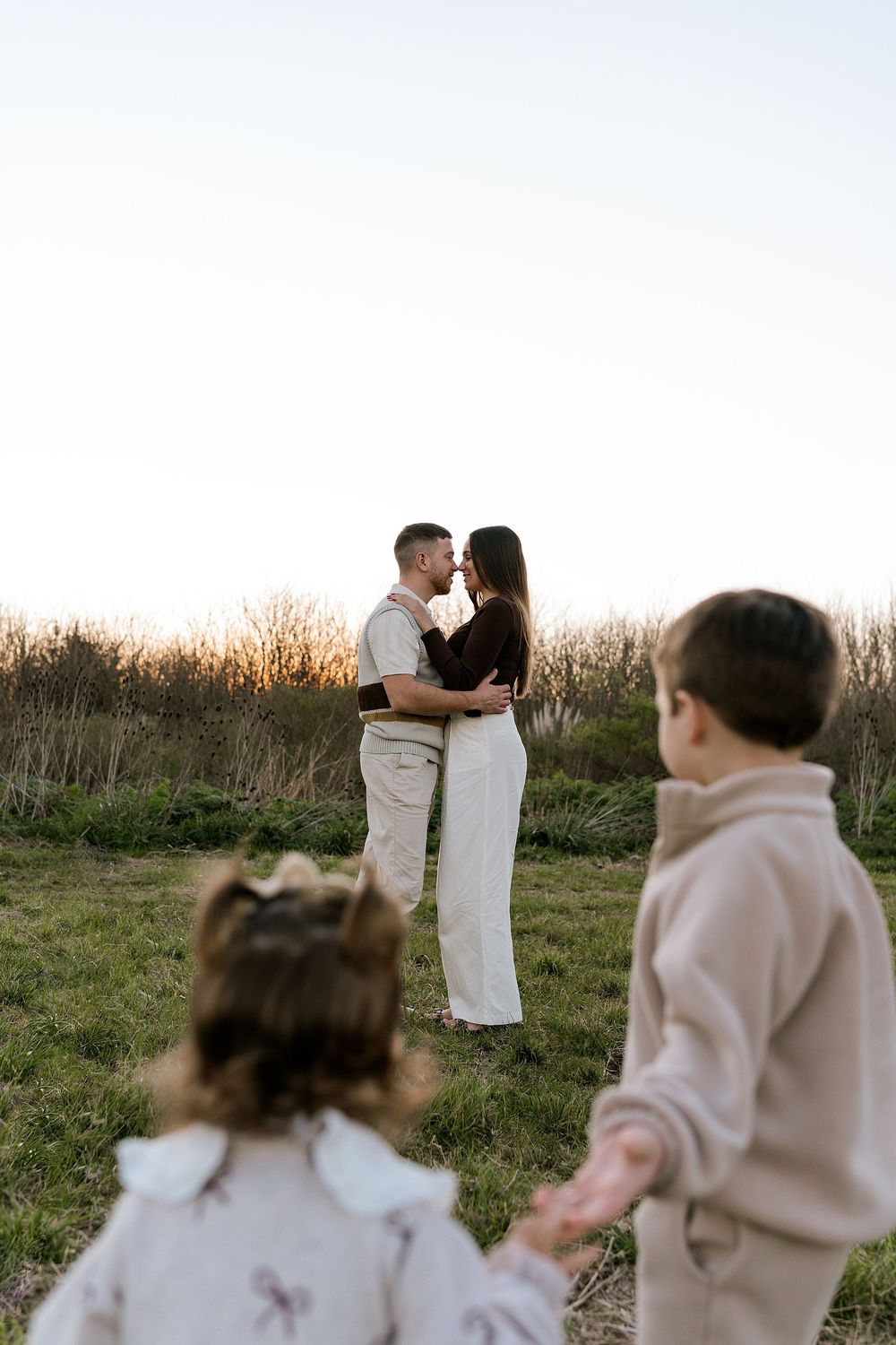 Romantic couple embraces in a golden field at sunset while young children watch from behind.