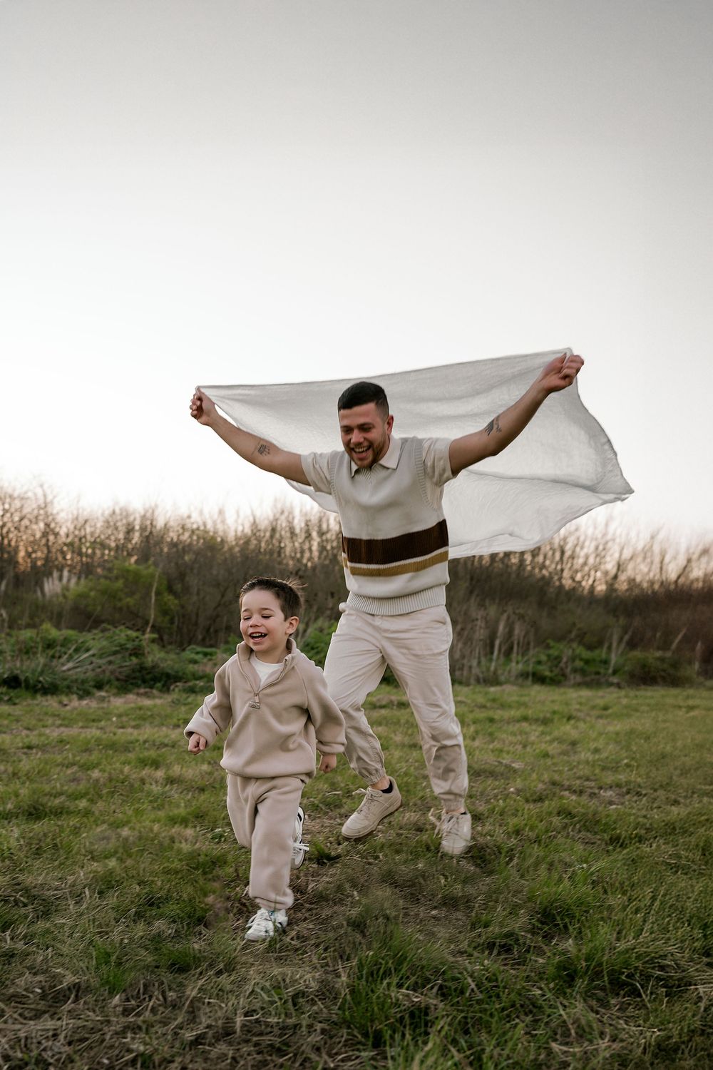 Two people in white clothing playfully jump and dance together on a grassy field at sunset.