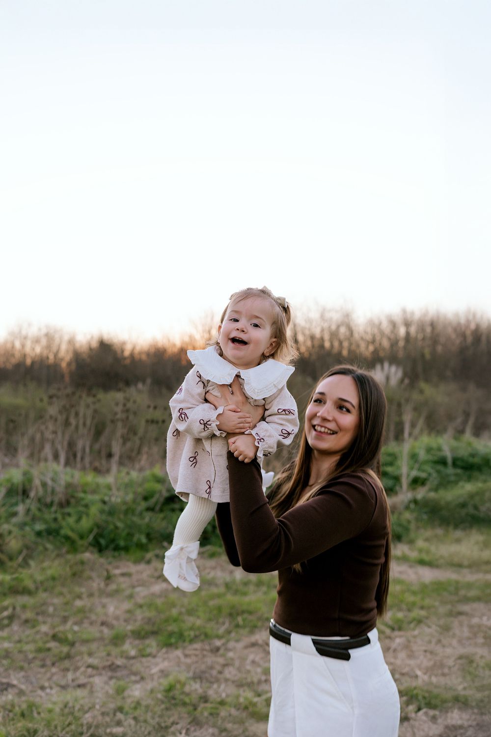 A person lifts and embraces a baby while laughing joyfully in an outdoor field during golden hour.