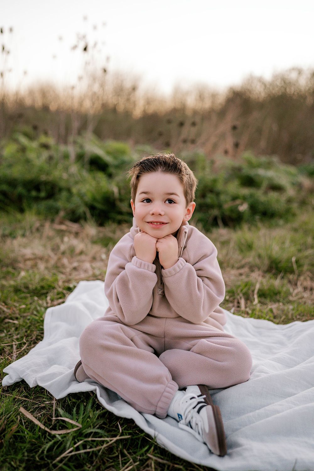 Child in pink sweater sits on white blanket outdoors while resting chin on hand in three similar poses.