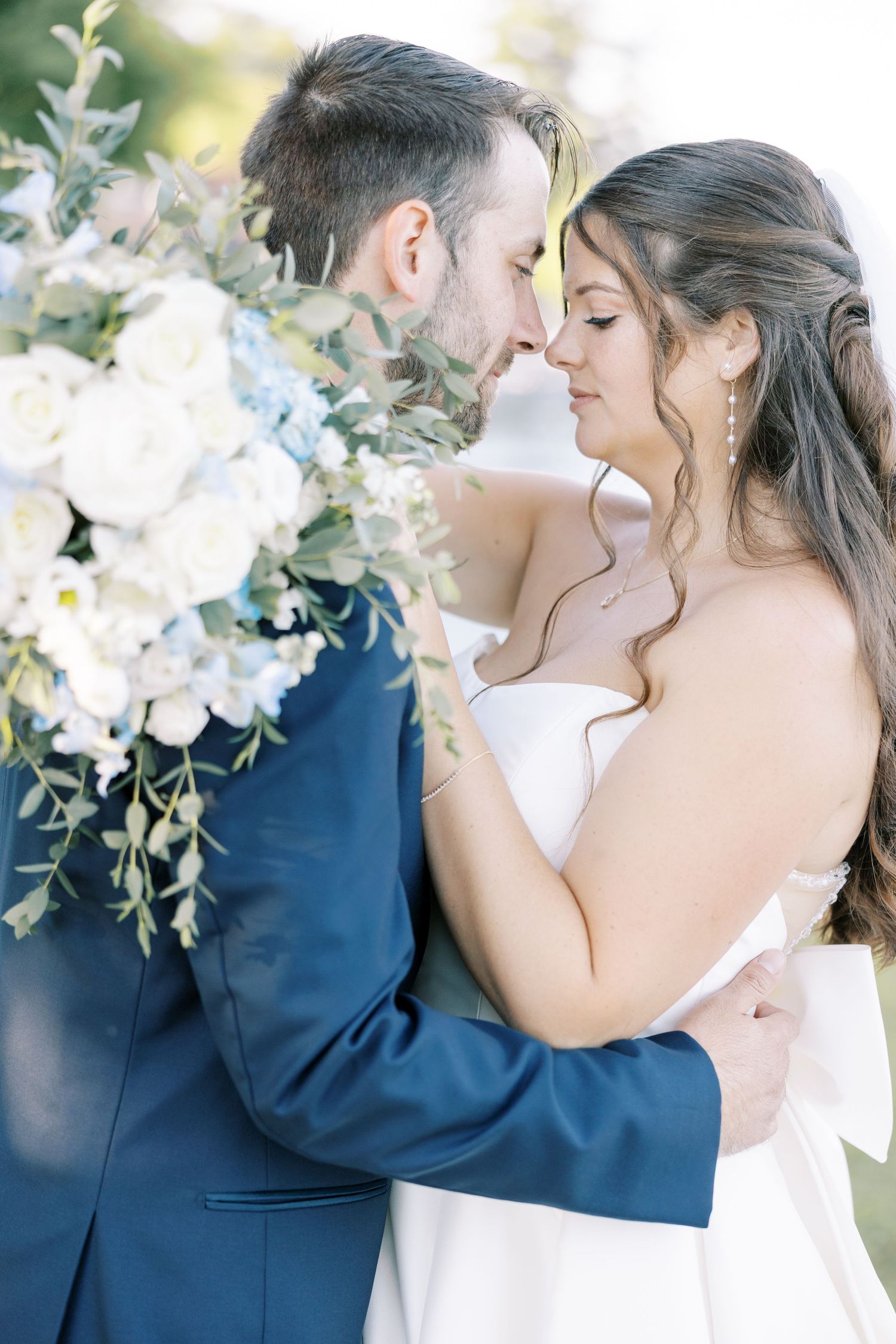 Romantic wedding photo with white floral arrangements as couple embraces outdoors in navy suit and white gown.