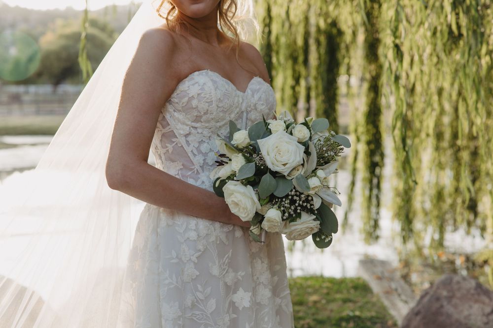 A person in a white wedding dress holds a bouquet of white flowers next to a willow tree by the water.