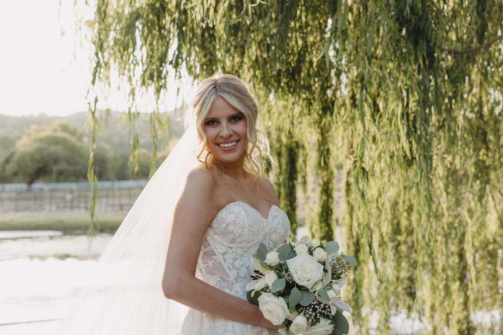 Bride in a white lace wedding dress holds a white floral bouquet while standing under cascading willow branches.