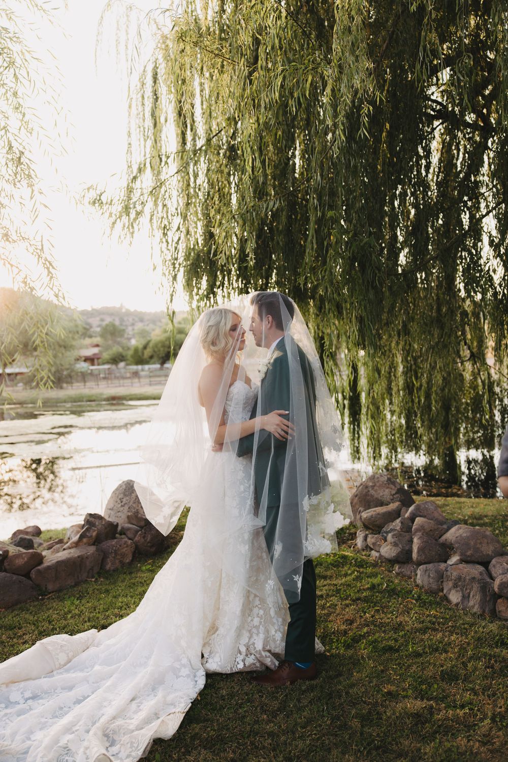 A wedding couple shares a romantic moment under a weeping willow tree by a pond at sunset.