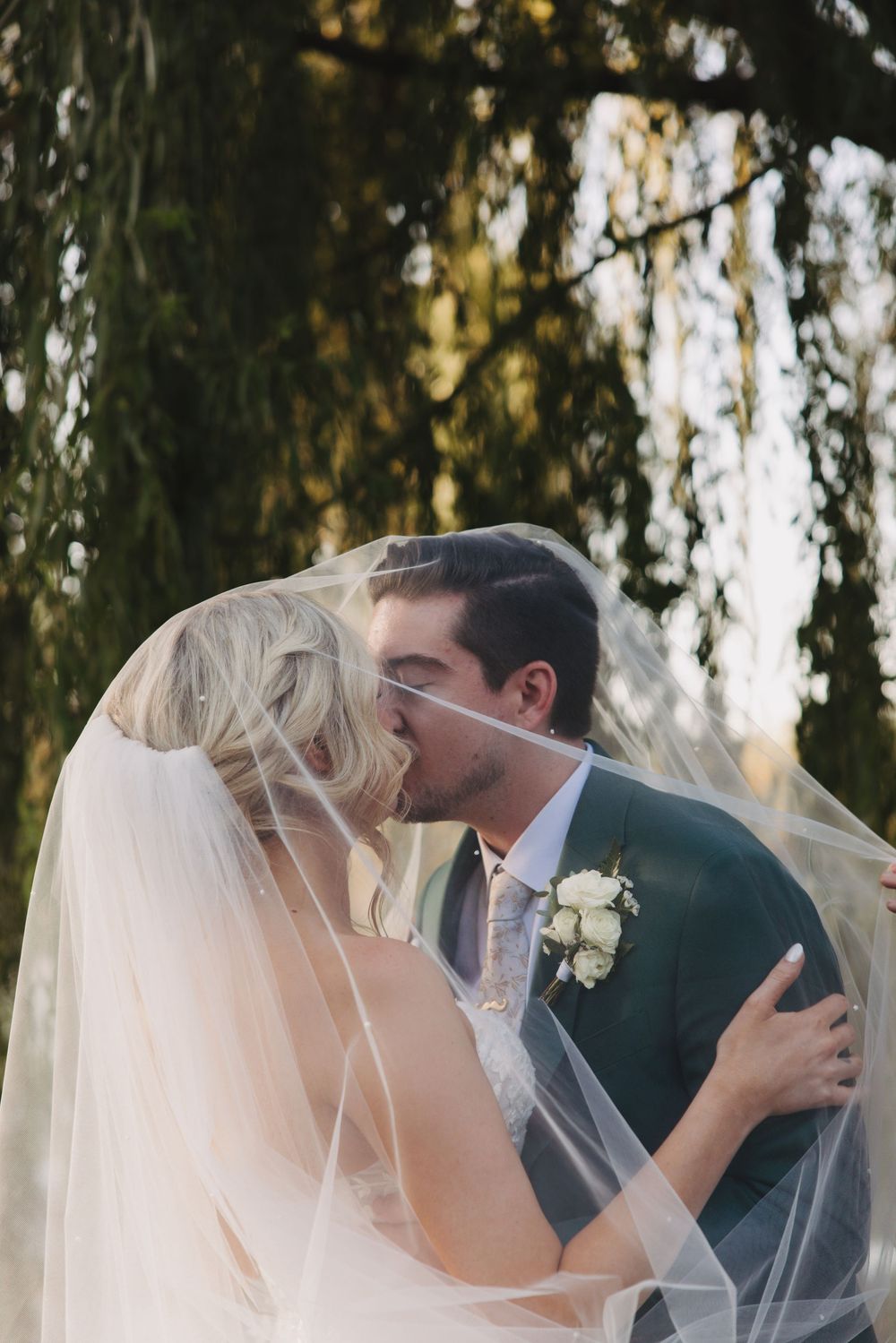 A romantic kiss shared between newlyweds under a drooping willow tree at sunset.