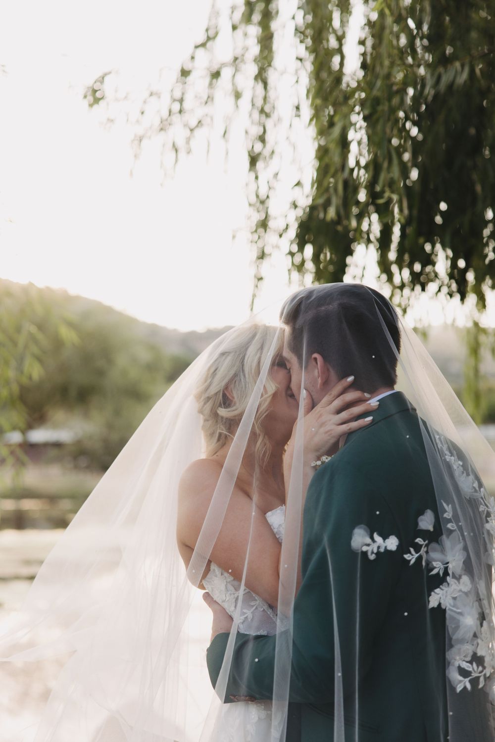 A couple in wedding attire share an intimate kiss under a flowing veil beside a willow tree.