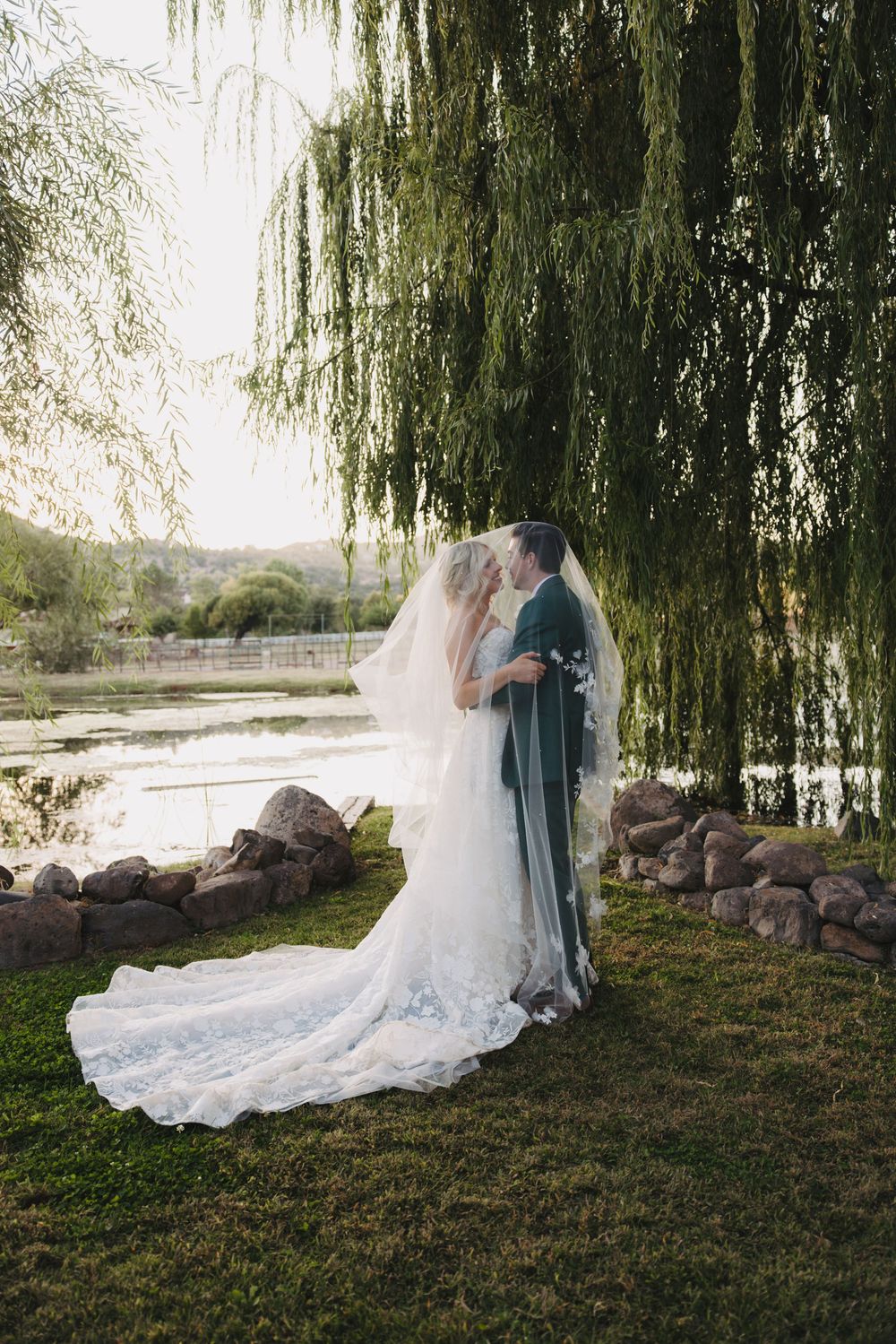 A flowing white wedding dress train trails across green grass beneath a weeping willow tree.
