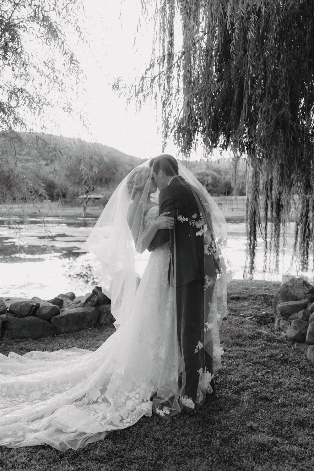 A romantic wedding couple embraces under a weeping willow tree by a serene lake with their reflection visible in the water.