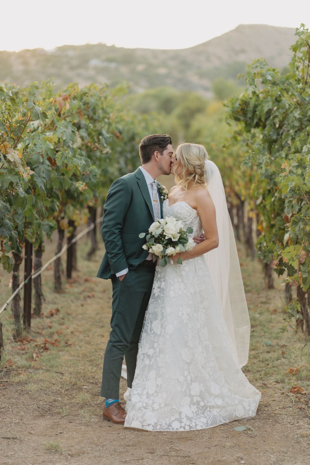 Newlyweds share a romantic kiss in a scenic vineyard setting on their wedding day.