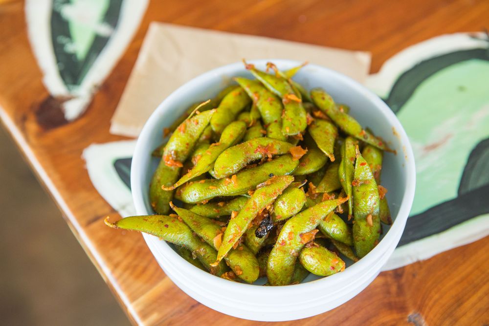 A bowl of grilled or roasted edamame beans with charred spots sits on a wooden table.